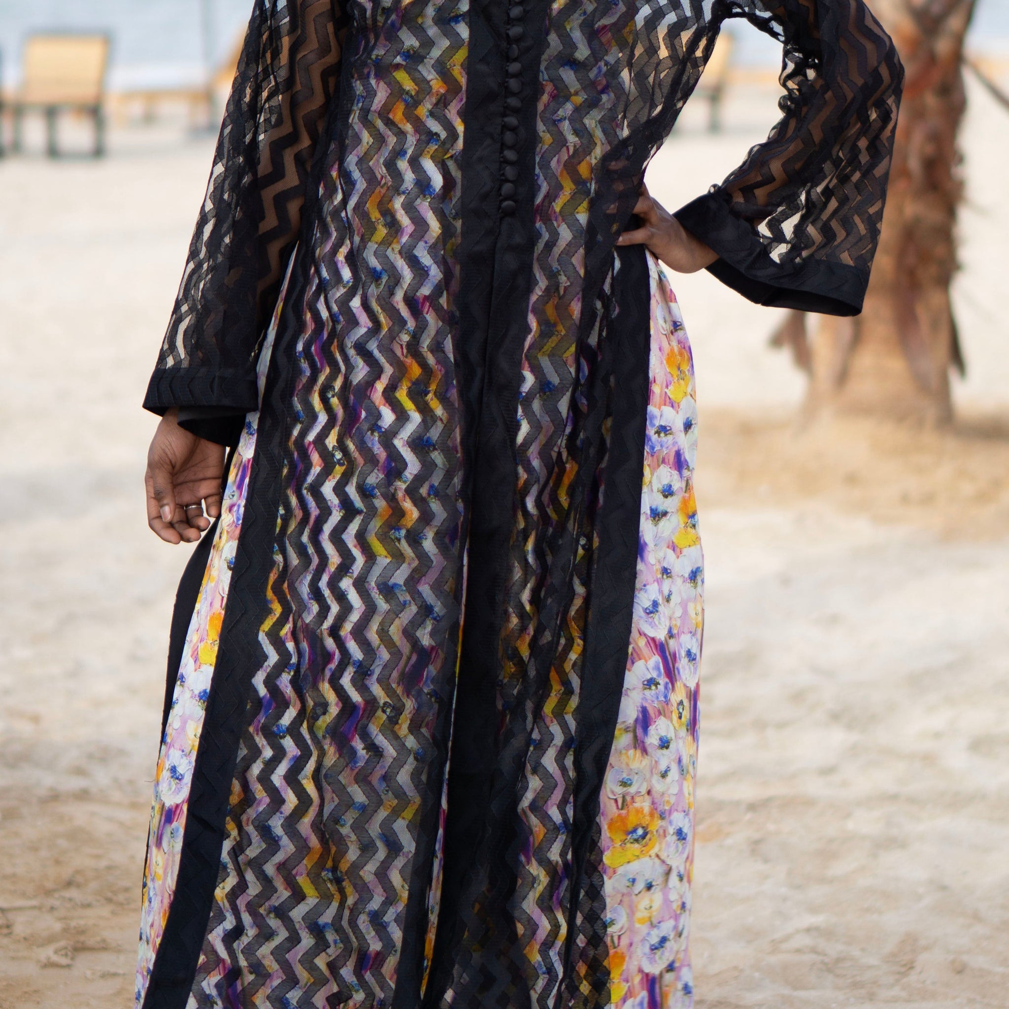 Long sheer black cover-up with a zigzag pattern with full-length sleeves and a bold chevron pattern. Features a lightweight, flowy design with a black button-down front closure, perfect for layering over swimwear or resort outfits. Styled over a floral dress on a beach.