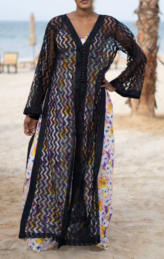 Long sheer black cover-up with a zigzag pattern with full-length sleeves and a bold chevron pattern. Features a lightweight, flowy design with a black button-down front closure, perfect for layering over swimwear or resort outfits. Styled over a floral dress on a beach.
