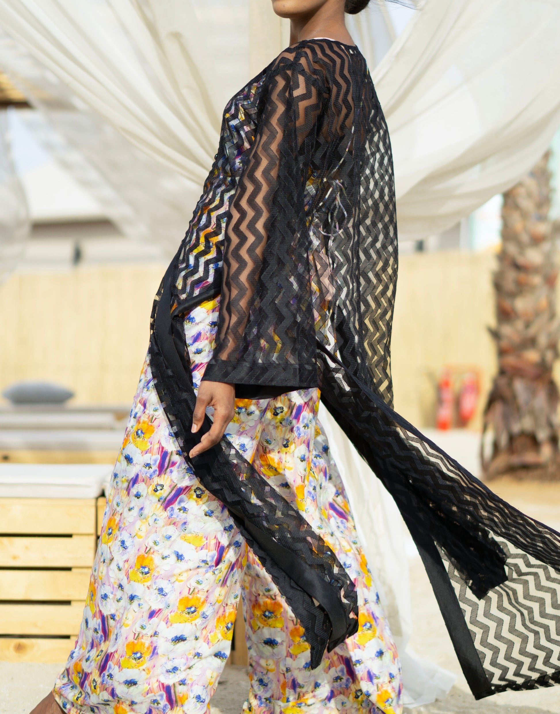 Long sheer black cover-up with a zigzag pattern with full-length sleeves and a bold chevron pattern. Features a lightweight, flowy design with a black button-down front closure, perfect for layering over swimwear or resort outfits. Styled over a floral dress on a beach.