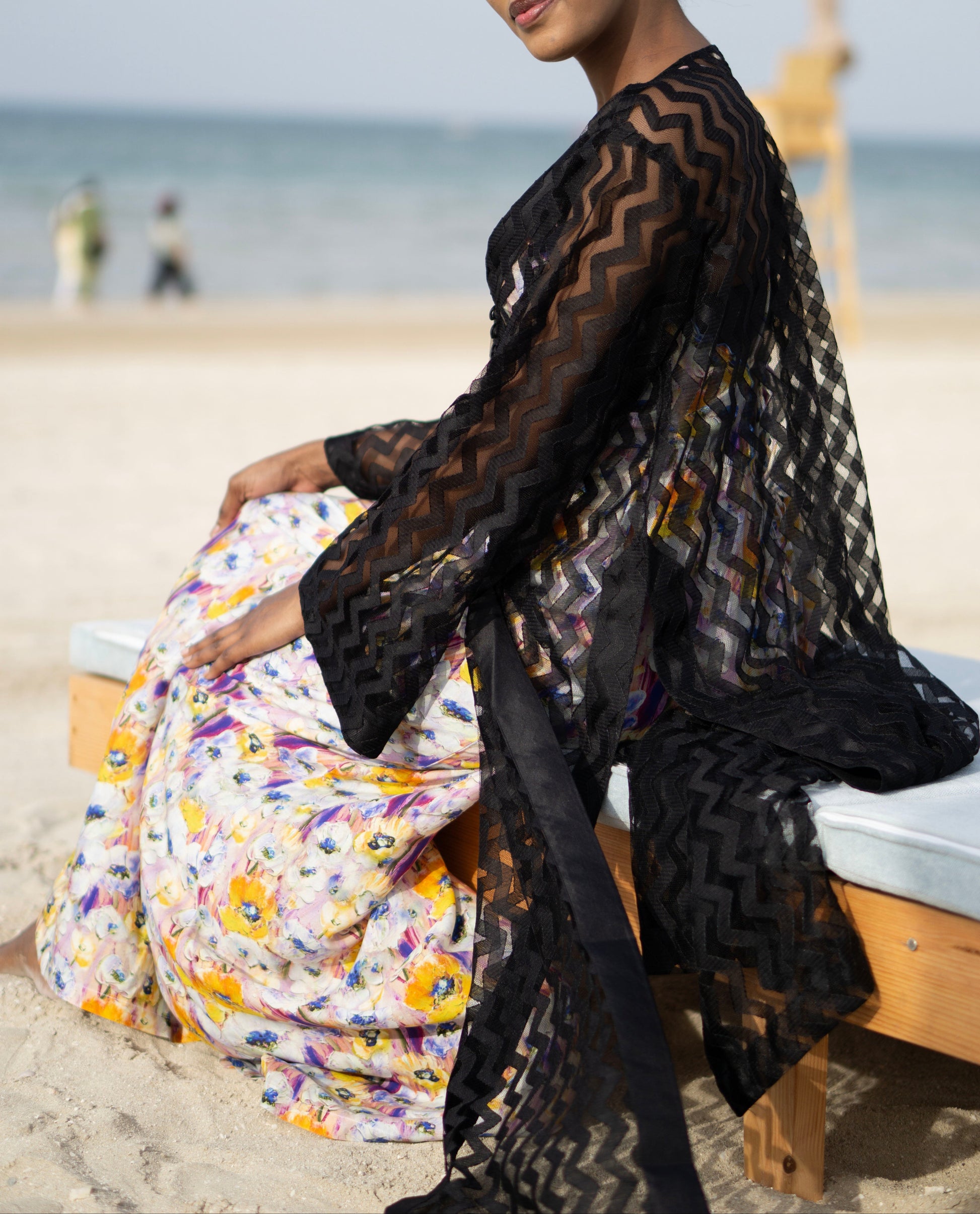 Long sheer black cover-up with a zigzag pattern with full-length sleeves and a bold chevron pattern. Features a lightweight, flowy design with a black button-down front closure, perfect for layering over swimwear or resort outfits. Styled over a floral dress on a beach.