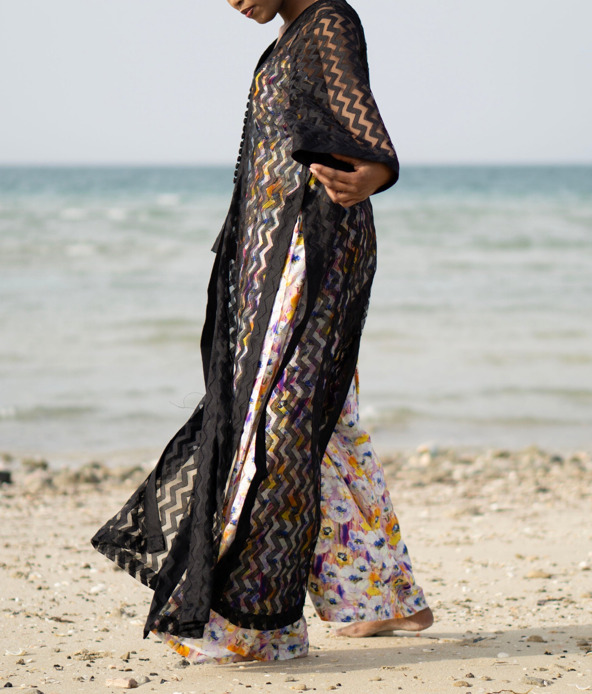 Long sheer black cover-up with a zigzag pattern with full-length sleeves and a bold chevron pattern. Features a lightweight, flowy design with a black button-down front closure, perfect for layering over swimwear or resort outfits. Styled over a floral dress on a beach.