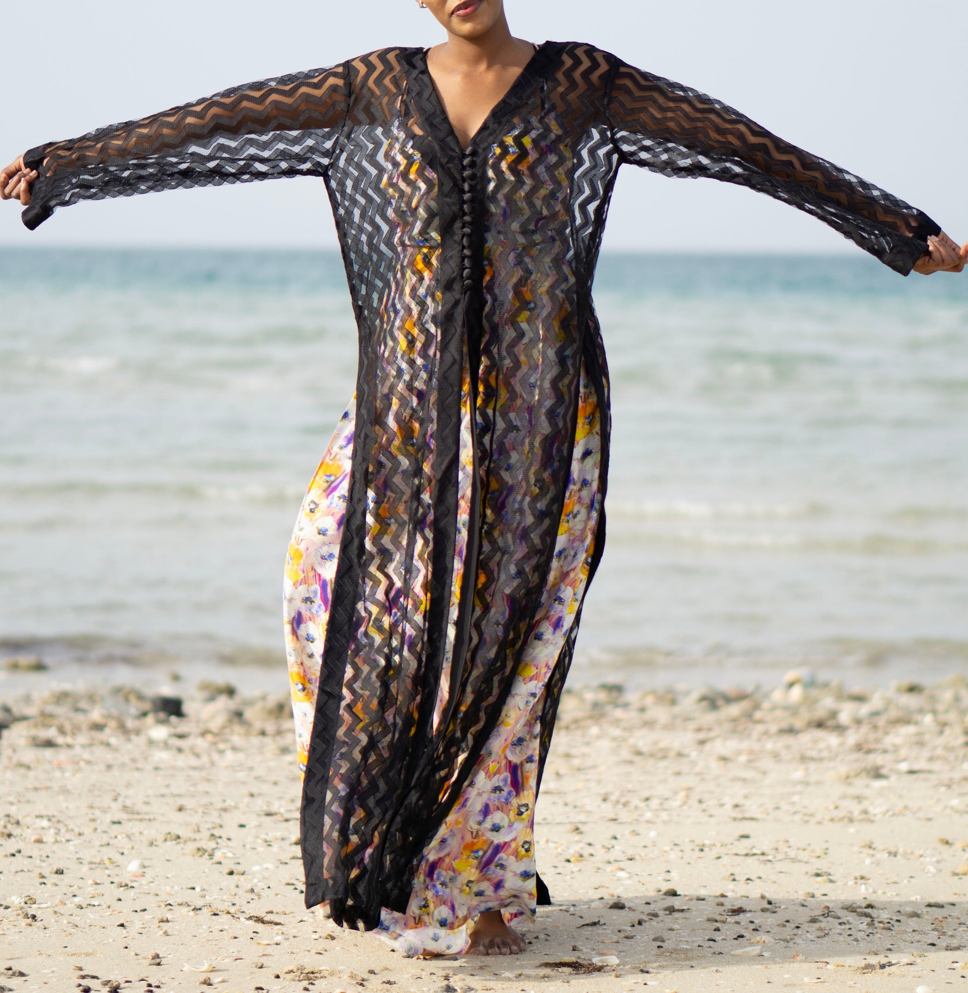 Long sheer black cover-up with a zigzag pattern with full-length sleeves and a bold chevron pattern. Features a lightweight, flowy design with a black button-down front closure, perfect for layering over swimwear or resort outfits. Styled over a floral dress on a beach.