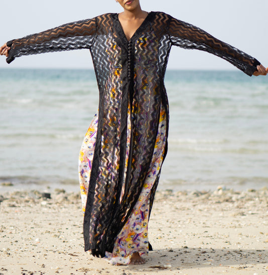 Long sheer black cover-up with a zigzag pattern with full-length sleeves and a bold chevron pattern. Features a lightweight, flowy design with a black button-down front closure, perfect for layering over swimwear or resort outfits. Styled over a floral dress on a beach.
