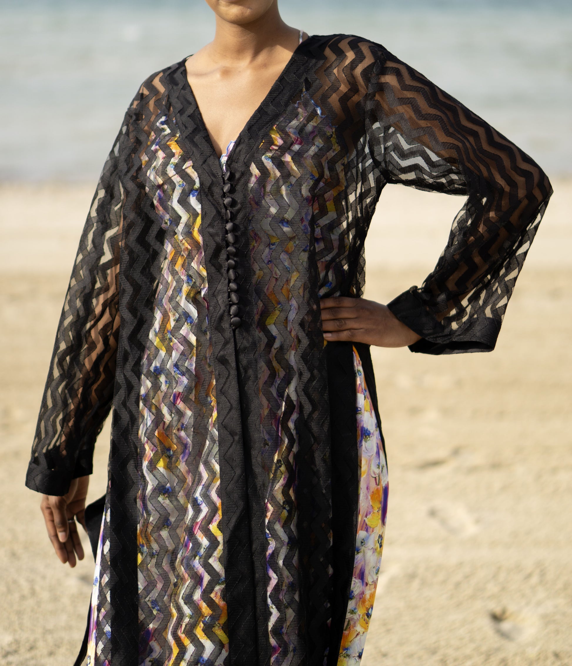 Long sheer black cover-up with a zigzag pattern with full-length sleeves and a bold chevron pattern. Features a lightweight, flowy design with a black button-down front closure, perfect for layering over swimwear or resort outfits. Styled over a floral dress on a beach.