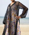Long sheer black cover-up with a zigzag pattern with full-length sleeves and a bold chevron pattern. Features a lightweight, flowy design with a black button-down front closure, perfect for layering over swimwear or resort outfits. Styled over a floral dress on a beach.