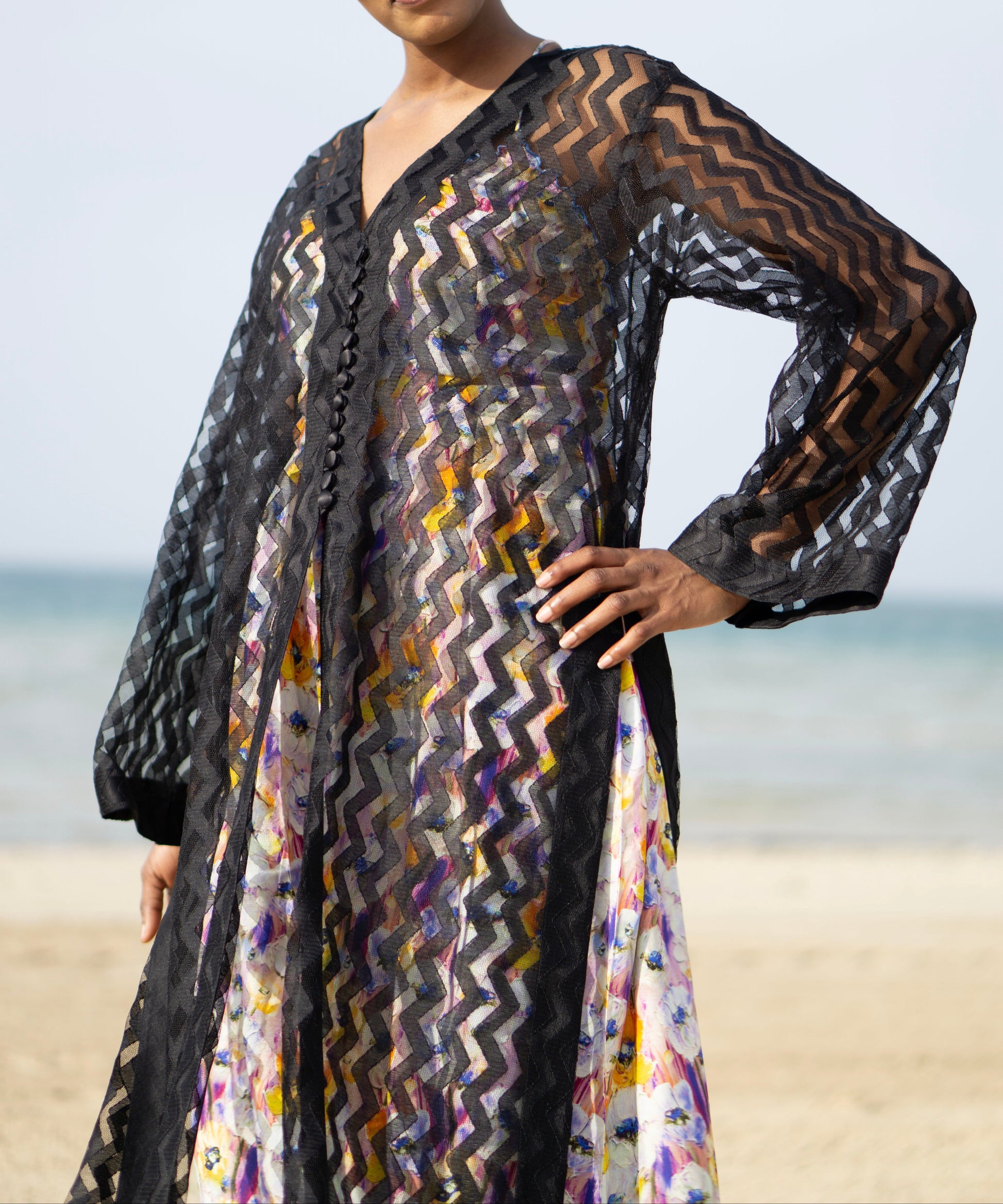 Long sheer black cover-up with a zigzag pattern with full-length sleeves and a bold chevron pattern. Features a lightweight, flowy design with a black button-down front closure, perfect for layering over swimwear or resort outfits. Styled over a floral dress on a beach.